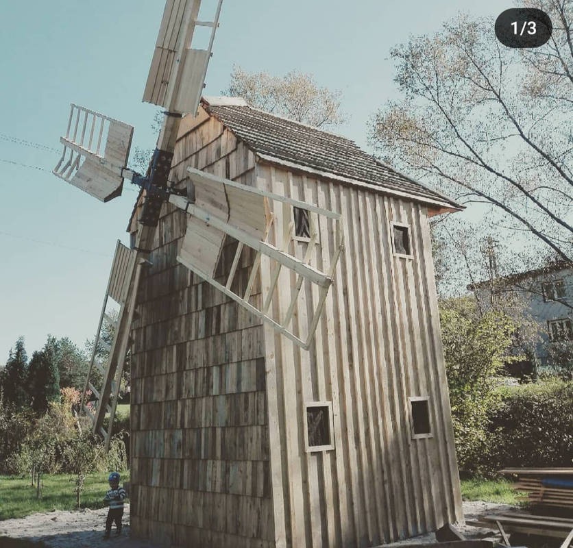 Nowy drewniany wiatrak z nieobrobionego drewna, częściowo pokryty gontem, z widocznym fragmentem ogrodu i małym dzieckiem w tle.
