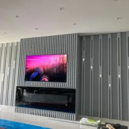 Ściana z dekoracyjnymi panelami w odcieniach szarości, z wbudowanym telewizorem i kominkiem elektrycznym, częściowo zabezpieczona folią ochronną na podłodze.