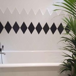 GAB Home Improvements - Biała wanna wolnostojąca z chromowaną armaturą na tle ściany z białych płytek ułożonych w romby z czarnymi akcentami, uzupełniona zieloną rośliną doniczkową.