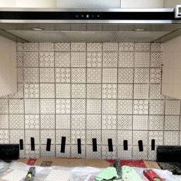 GAB Home Improvements - Montaż płytek dekoracyjnych w kuchni, widoczne czarne kliny dystansowe pod płytkami, okap kuchenny z timerem, fragment szafki.