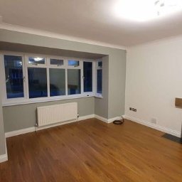 GAB Home Improvements - Jasne, puste pomieszczenie po remoncie z drewnianą podłogą, grzejnikiem pod oknem wykuszowym i kominkiem z drewnianą belką.