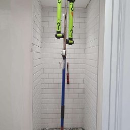 GAB Home Improvements - Wąska kabina prysznicowa w trakcie kładzenia białych płytek, użycie narzędzia do podtrzymywania płytek, widoczna podłoga w trakcie prac remontowych.