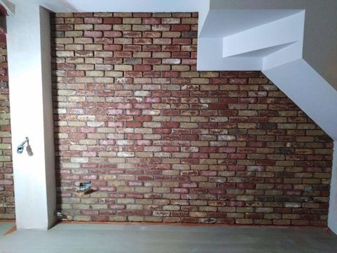 Ściana z cegły klinkierowej w odcieniach czerwieni i beżu, z widoczną instalacją wodną wystającą ze ściany i nietypowym, schodkowym elementem dekoracyjnym na suficie.