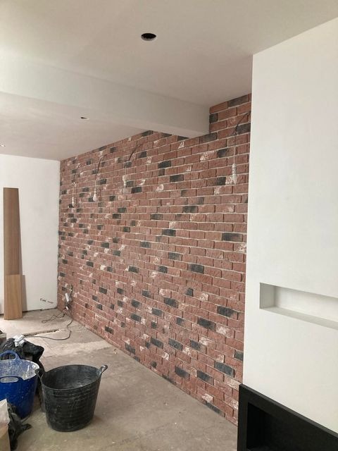 Ściana wyłożona cegłą dekoracyjną w trakcie prac wykończeniowych, widoczne wiadro i przewody, surowy stan podłogi.