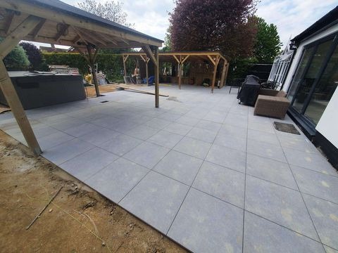 Nowa, jasnoszara, kwadratowa nawierzchnia tarasu z widocznymi drewnianymi konstrukcjami altan, jacuzzi i grilla w tle, częściowo wykończona, z widocznymi pozostałościami prac ziemnych.