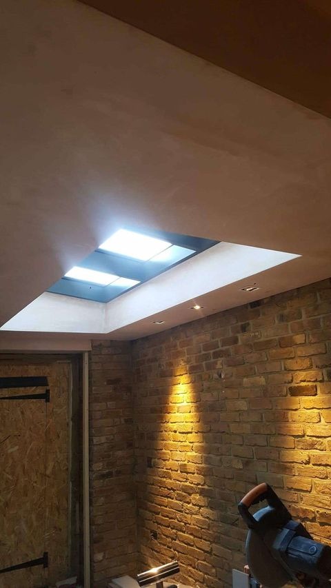 Wnętrze w trakcie prac wykończeniowych: surowa ceglana ściana, osadzony świetlik w suficie z widocznymi lampami i szpachlowaną powierzchnią, fragment drzwi z surowej płyty oraz narzędzia na podłodze.