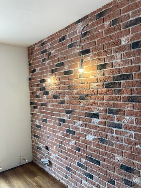 Ściana z cegły klinkierowej w odcieniach czerwieni, czerni i bieli, oświetlona dwoma wiszącymi żarówkami na kablach, z widocznymi gniazdkami elektrycznymi i fragmentem drewnianej podłogi.