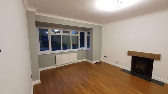 Jasne, puste pomieszczenie po remoncie z drewnianą podłogą, grzejnikiem pod oknem wykuszowym i kominkiem z drewnianą belką.