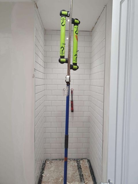 Wąska kabina prysznicowa w trakcie kładzenia białych płytek, użycie narzędzia do podtrzymywania płytek, widoczna podłoga w trakcie prac remontowych.