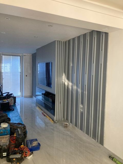 Montaż szarej, pionowej okładziny ściennej z wnęką na telewizor, widoczne narzędzia i poziomica na podłodze, w tle okno.