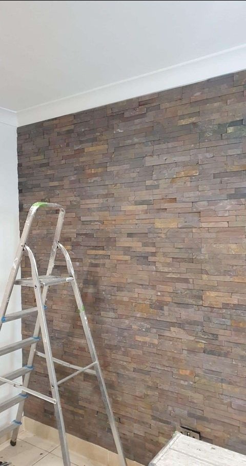 Ściana wykończona dekoracyjną okładziną kamienną w odcieniach brązu i beżu, widoczna aluminiowa drabina i fragment podłogi z drobnymi zabrudzeniami budowlanymi.