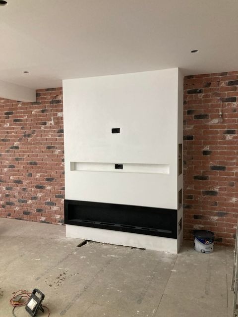 Ściana z cegły dekoracyjnej i biała zabudowa kominkowa z czarnym wkładem, w trakcie prac wykończeniowych w Grudziądzu, widoczny przewód elektryczny i puszka farby.