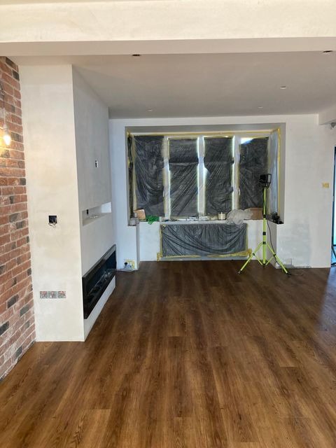 Salon w trakcie wykańczania: świeżo położona podłoga z paneli, ściana z imitacji cegły, okno zabezpieczone folią malarską, widoczny fragment kominka elektrycznego.