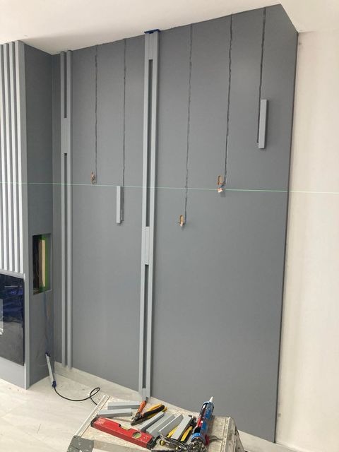 Montaż dekoracyjnych paneli ściennych w szarej tonacji, widoczne narzędzia i poziomica laserowa wskazująca linię, etap prac wykończeniowych.