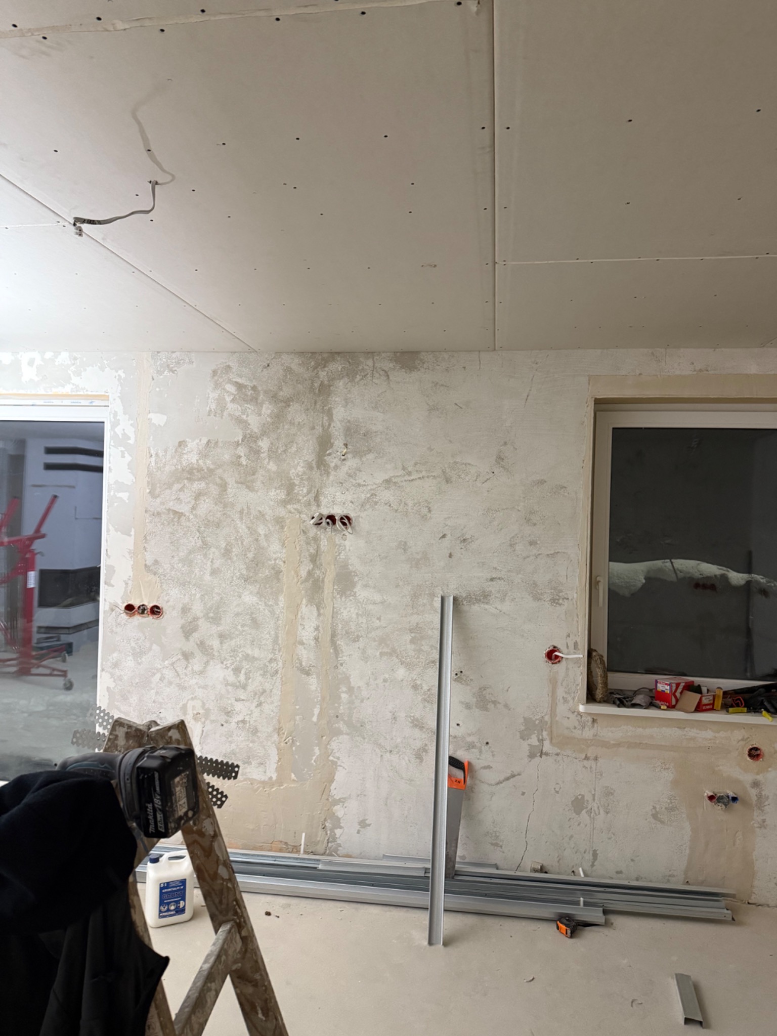 Ściana w trakcie wygładzania gipsem, widoczne puszki elektryczne, profile i narzędzia. Sufit z płyt kartonowo-gipsowych. Prace wykończeniowe w surowym wnętrzu.