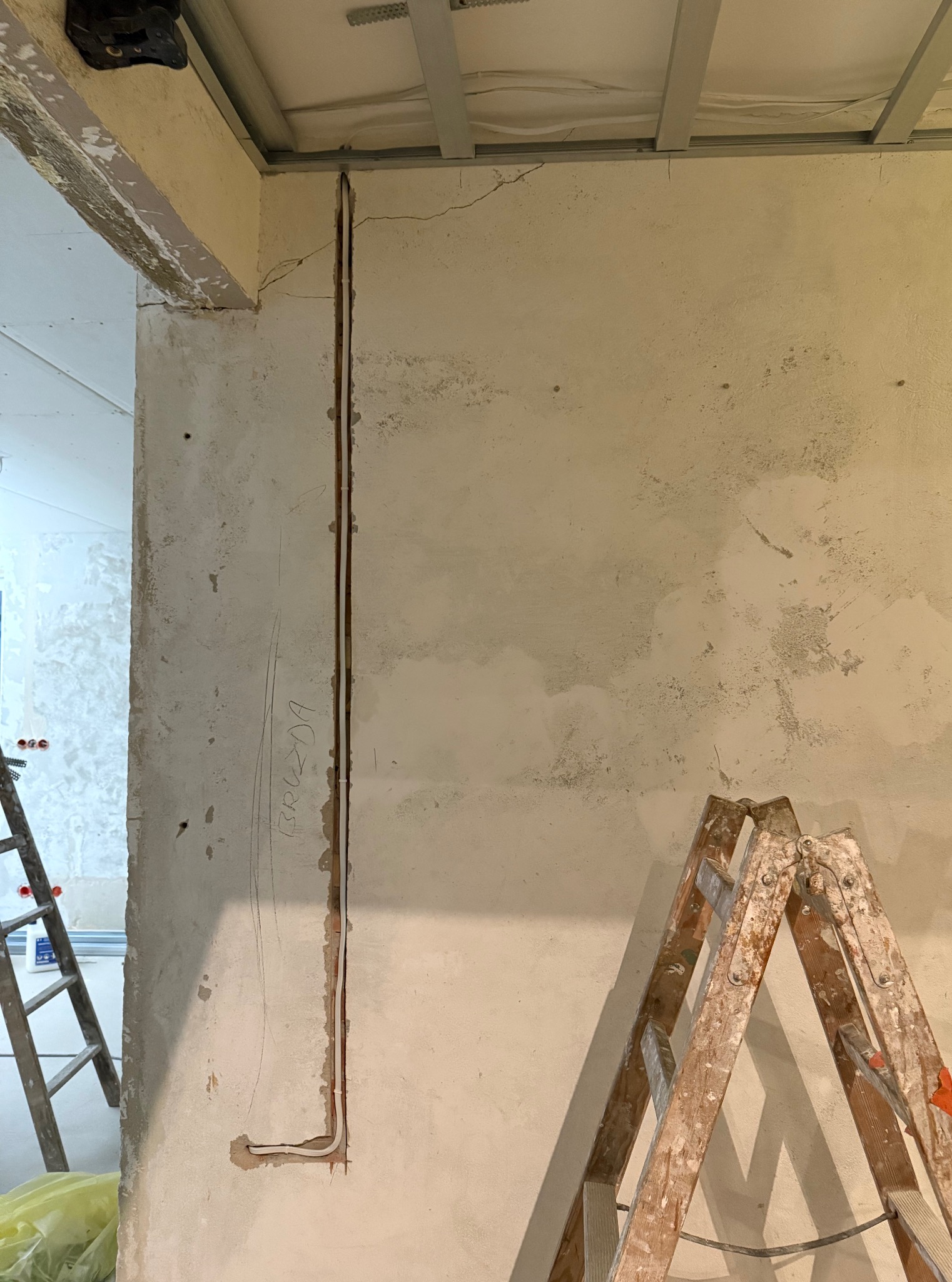 Bruzda z kablem w ścianie, przygotowana do instalacji elektrycznej. Widoczne drabiny malarskie i surowy stan wykończenia wnętrza. Ściana z napisem.