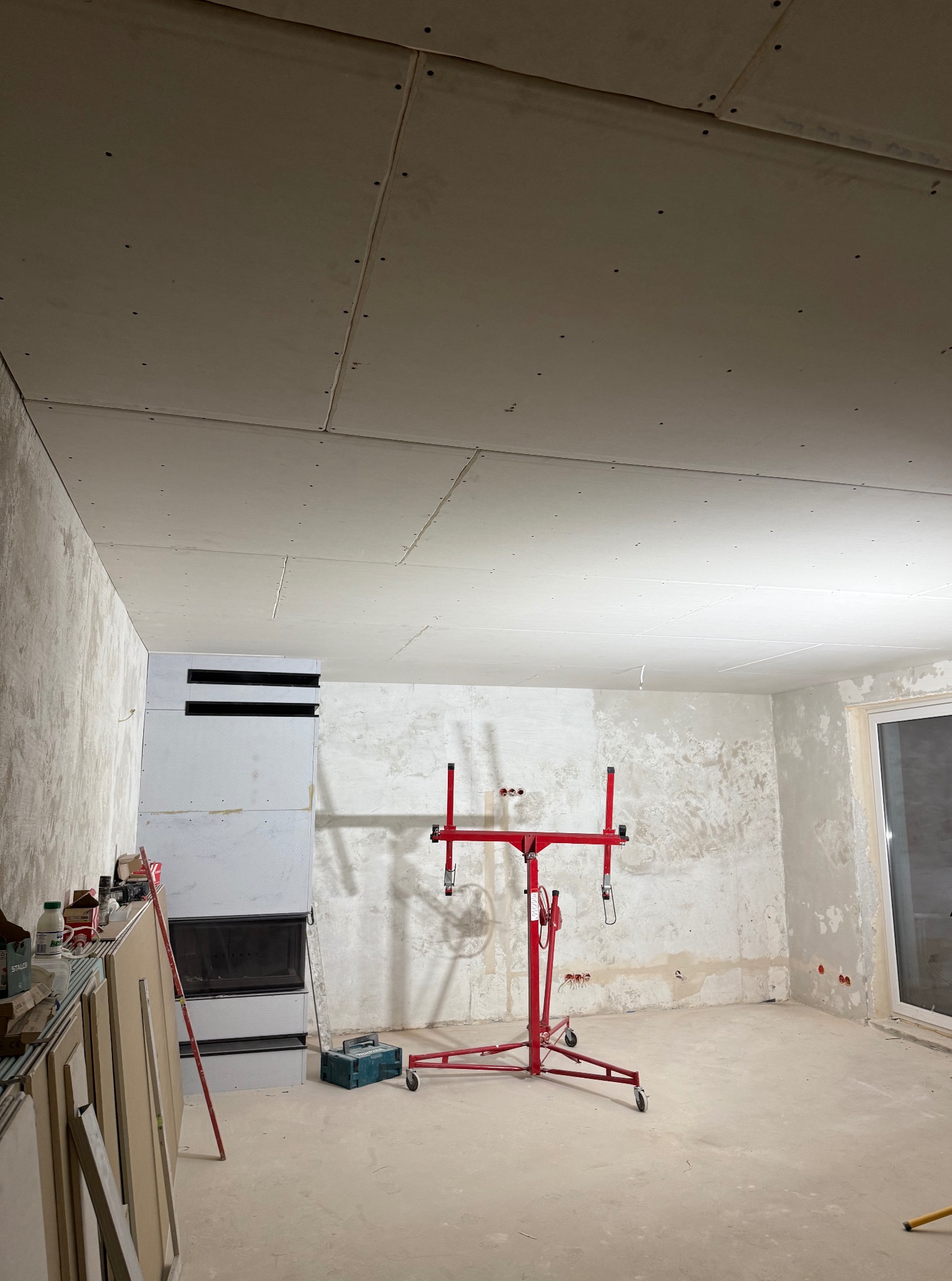 Montaż płyt gipsowo-kartonowych na suficie w surowym wnętrzu. Widoczny podnośnik do płyt i przygotowane ściany. Etap prac wykończeniowych.