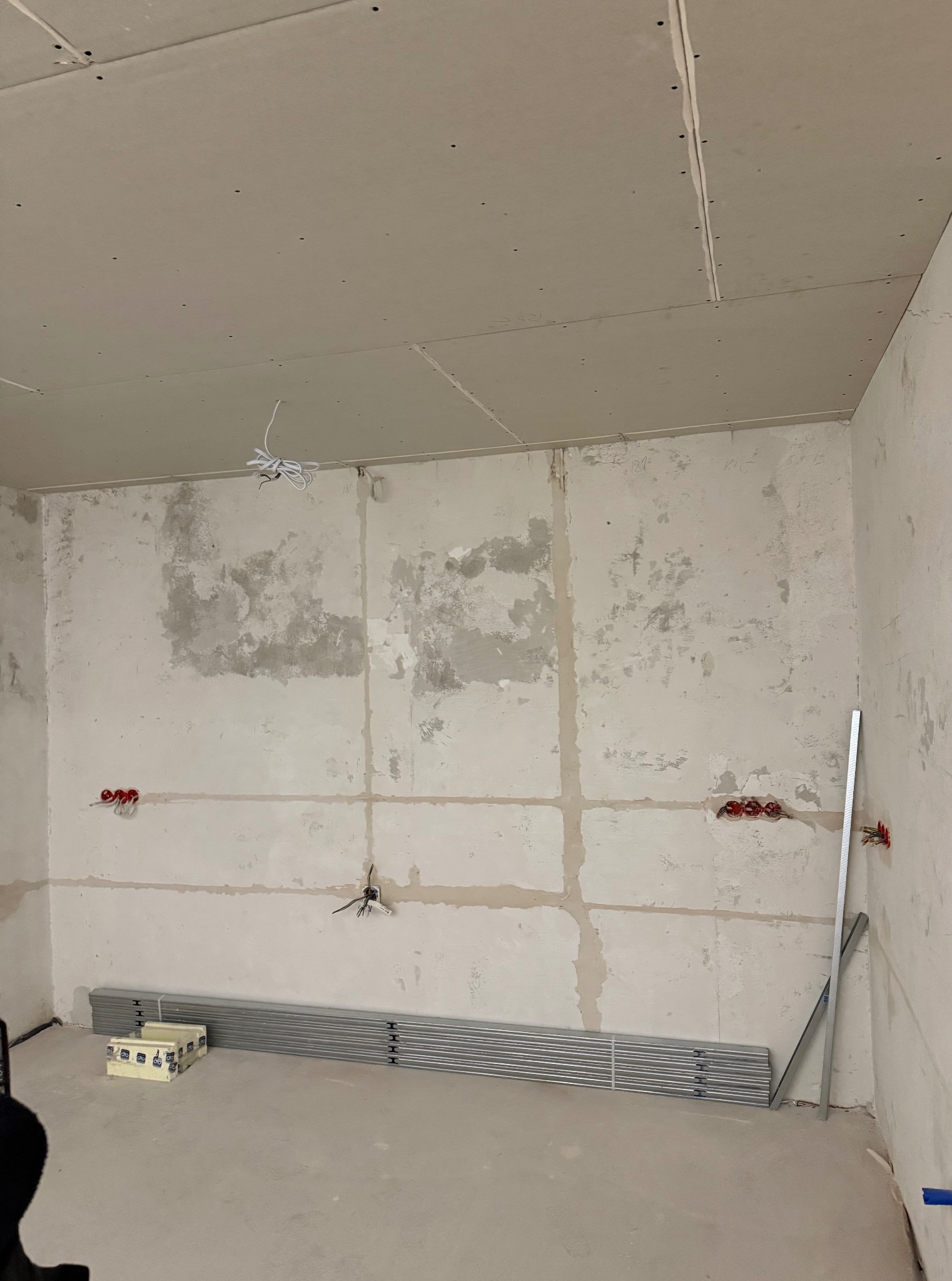 Surowy pokój w trakcie remontu: płyty gipsowe na suficie, ściany z widocznymi śladami szpachlowania, puszki elektryczne i profile na podłodze. Przygotowanie do wykończenia.