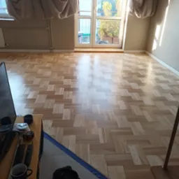 Jasne, nowo ułożone panele podłogowe w jodełkę, widok na balkon i okno z zasłonami, telewizor w rogu kadru