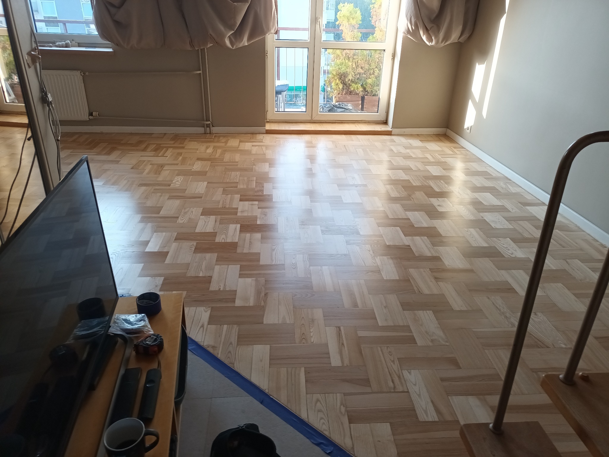 Jasne, nowo ułożone panele podłogowe w jodełkę, widok na balkon i okno z zasłonami, telewizor w rogu kadru