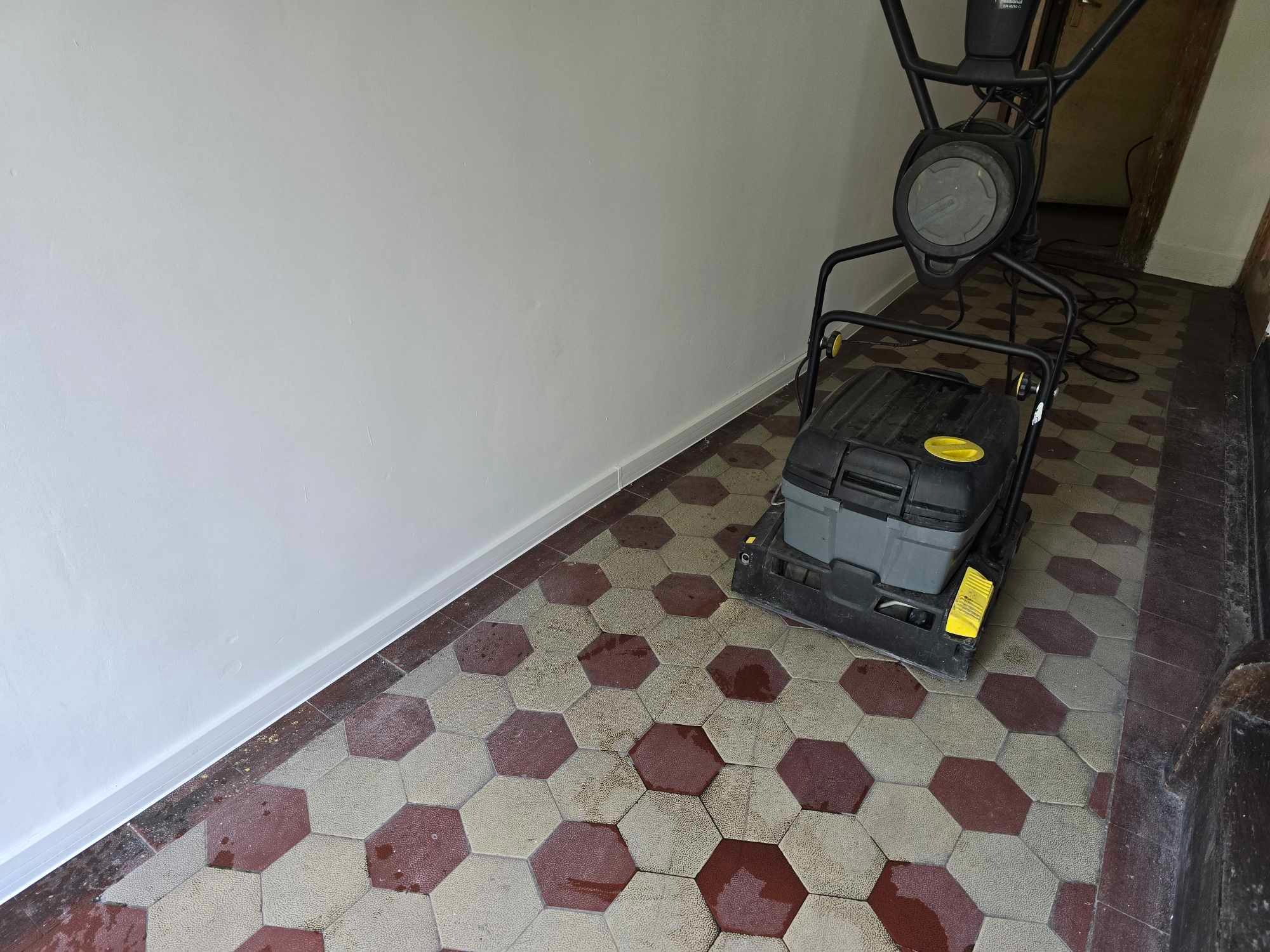 Maszyna do renowacji podłóg stoi na heksagonalnej posadzce z terakoty, częściowo odnowionej, z widocznymi śladami wilgoci i różnicami w kolorze między odnowionymi i nieodnowionymi fragmentami.