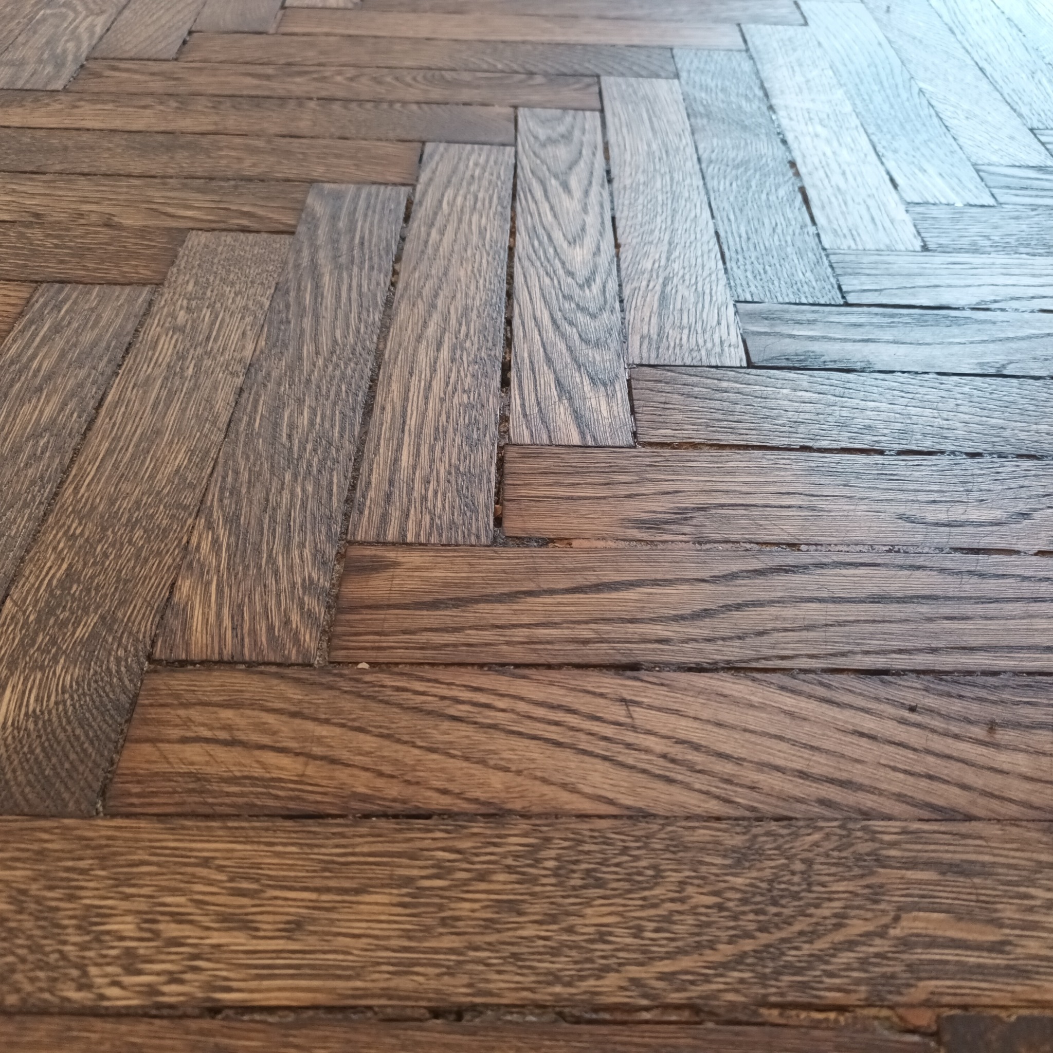 Ułożony parkiet w jodełkę o naturalnej barwie drewna z widocznymi słojami i drobnymi niedoskonałościami w spoinach.