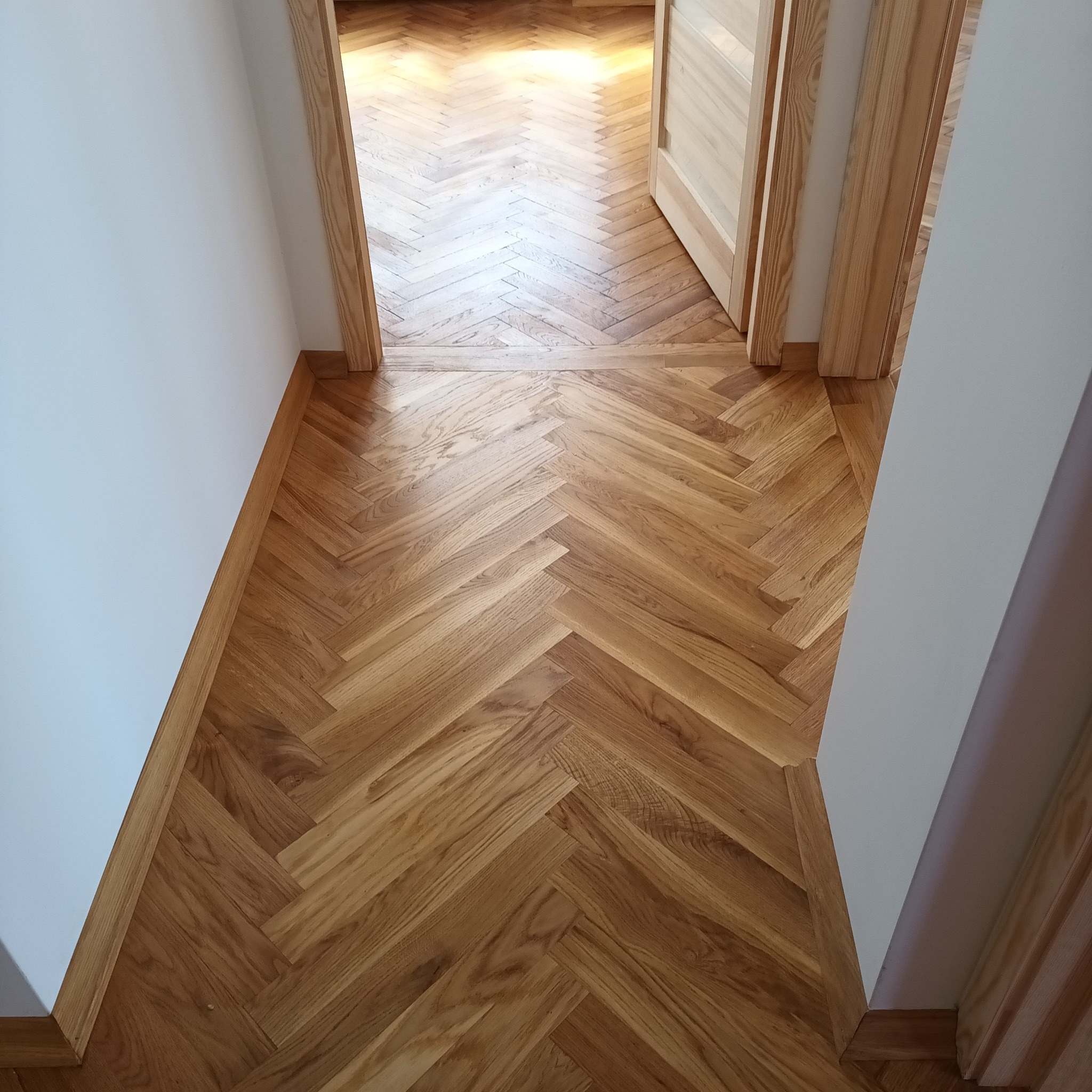 Ułożony parkiet w jodełkę w przejściu między pokojami, widoczne listwy przypodłogowe i ościeżnice z jasnego drewna.