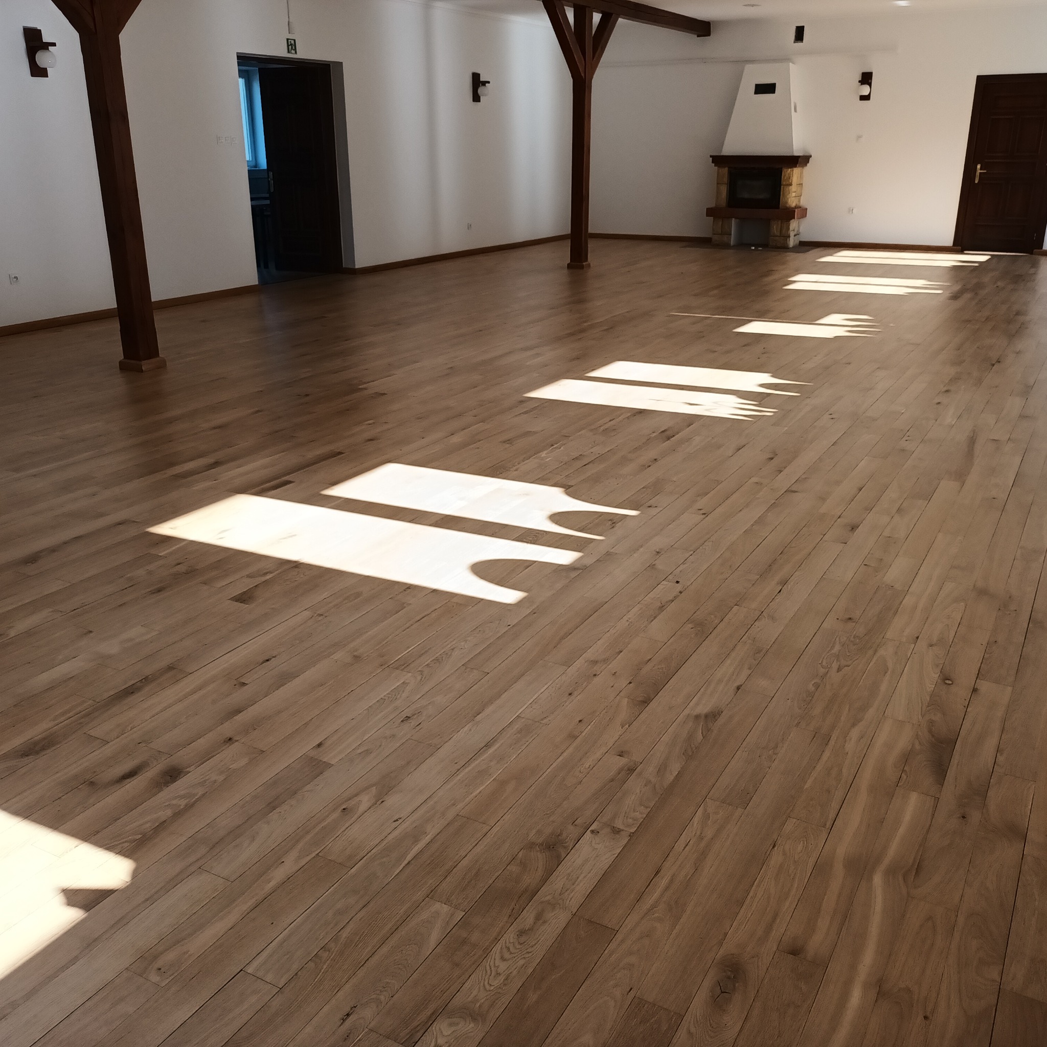 Jasny, wycyklinowany parkiet w Lidzbarku Warmińskim, z widocznymi promieniami słonecznymi tworzącymi geometryczne wzory na podłodze, w przestronnym wnętrzu z kominkiem.