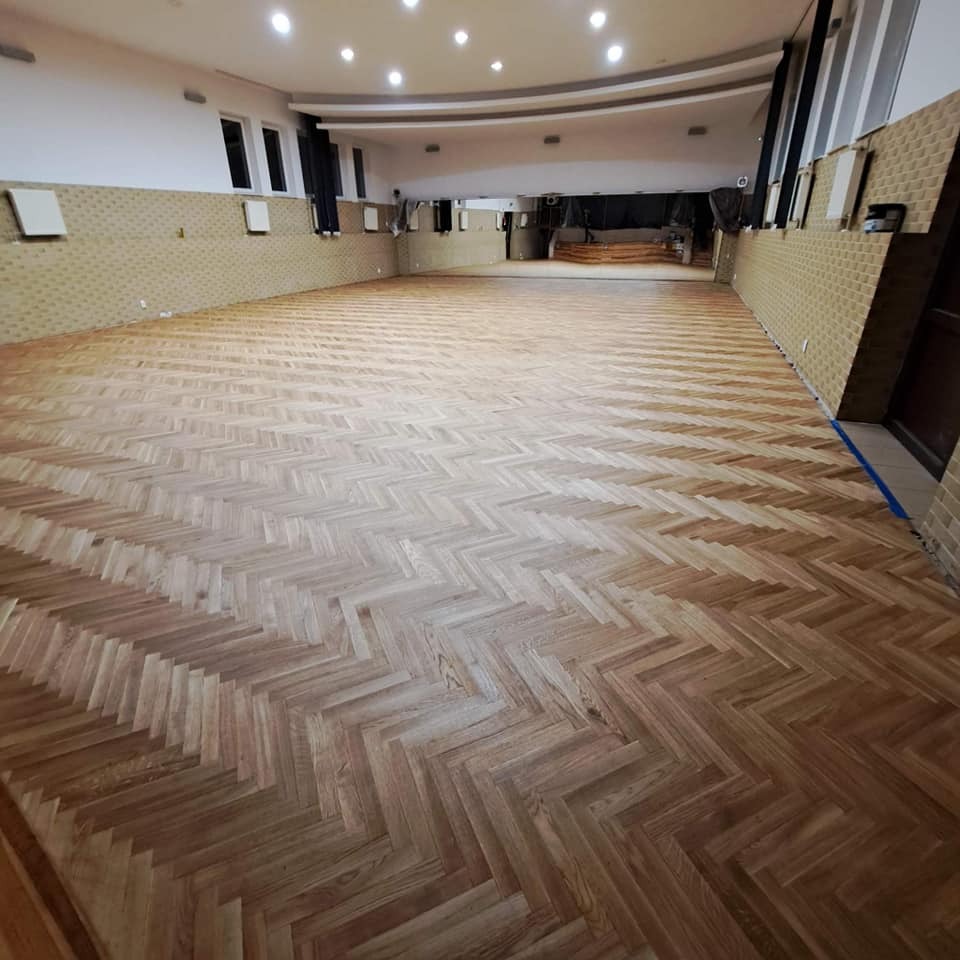 Ułożony parkiet w jodełkę francuską na dużej sali, widoczne panele ścienne i oświetlenie sufitowe.