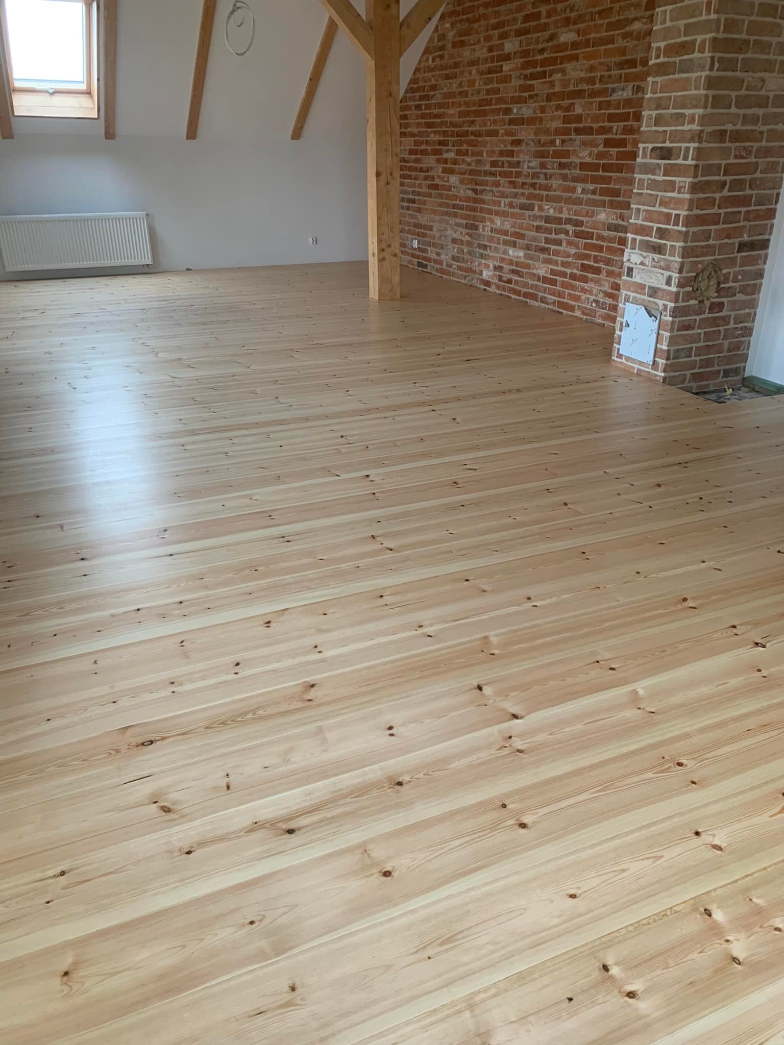Jasne, świeżo wycyklinowane deski podłogowe z widocznymi słojami drewna w przestronnym poddaszu z ceglaną ścianą i drewnianą konstrukcją nośną.