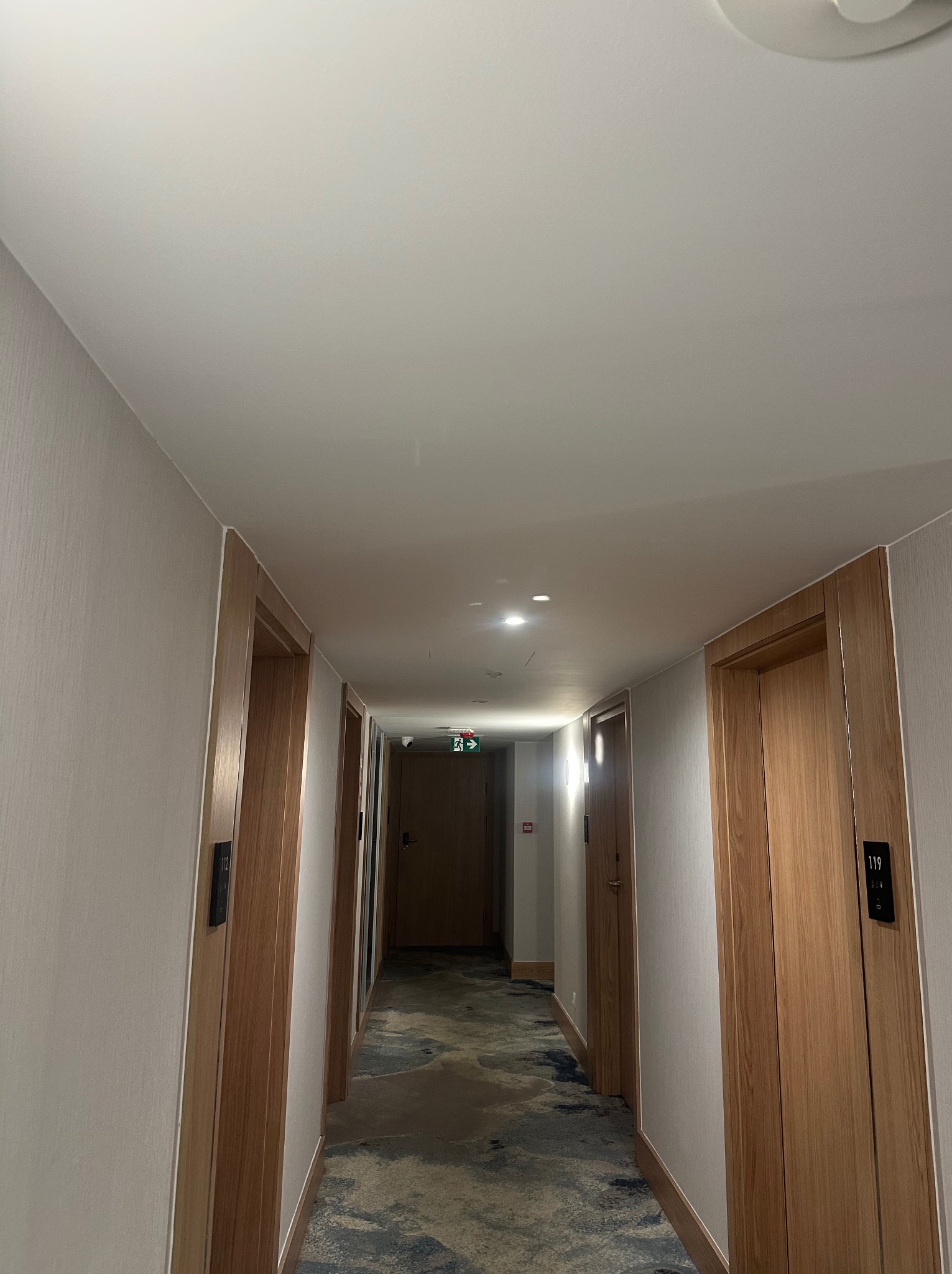 Długi korytarz hotelowy z wieloma drzwiami wykończonymi drewnem i numerami pokoi, podłoga pokryta wzorzystą wykładziną, oświetlenie sufitowe.
