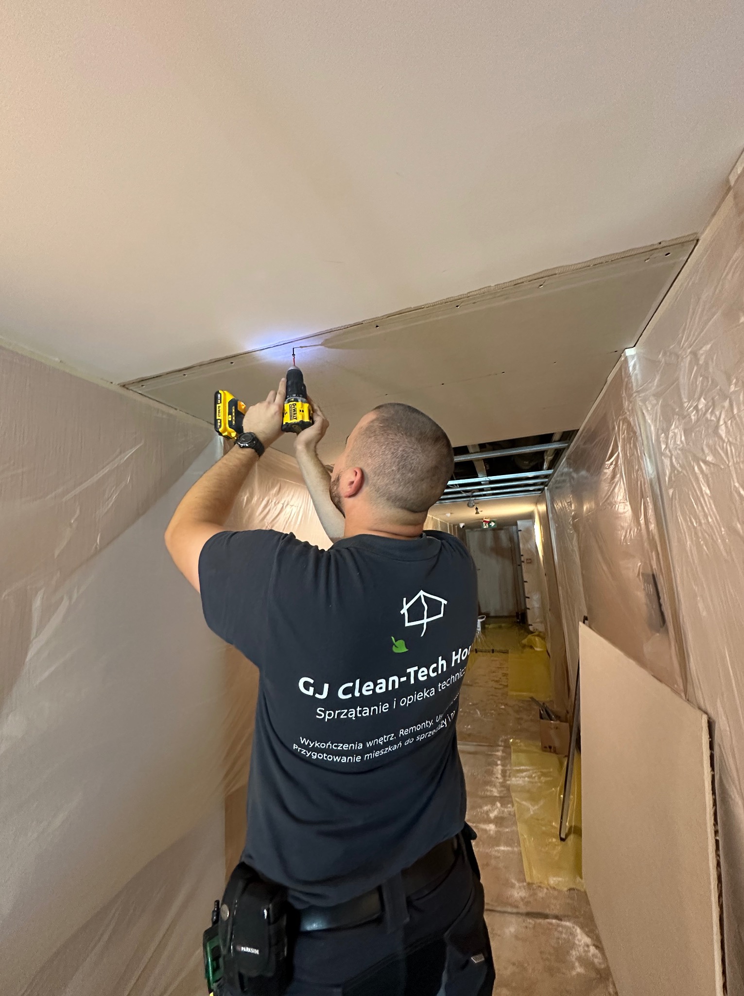 Pracownik wierci w suficie podwieszanym z płyty kartonowo-gipsowej, widoczny fragment rusztu, zabezpieczona folią przestrzeń, logo firmy na koszulce.