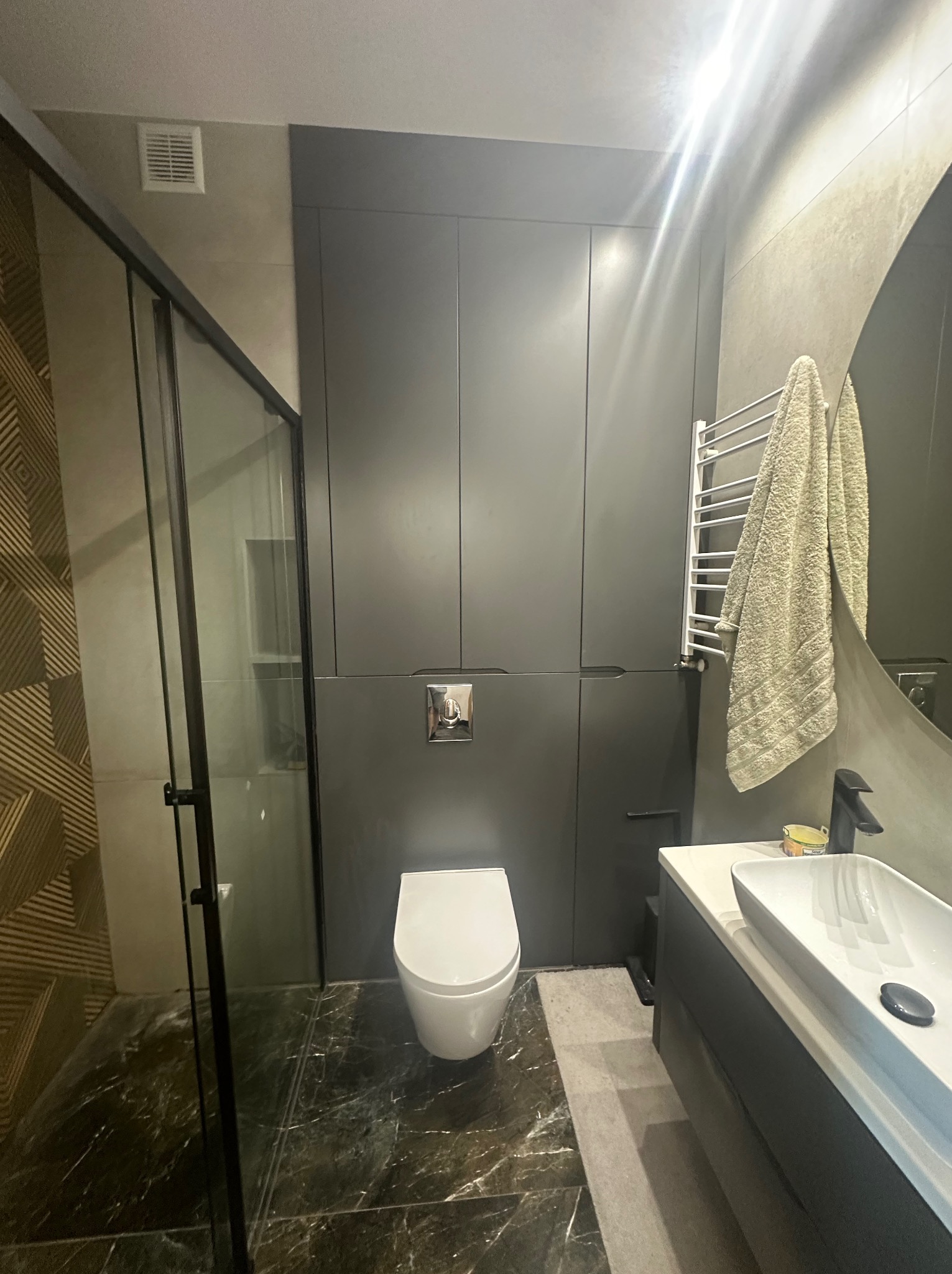 Nowoczesna łazienka z kabiną prysznicową typu walk-in, ciemną zabudową meblową, marmurową posadzką i okrągłym lustrem.