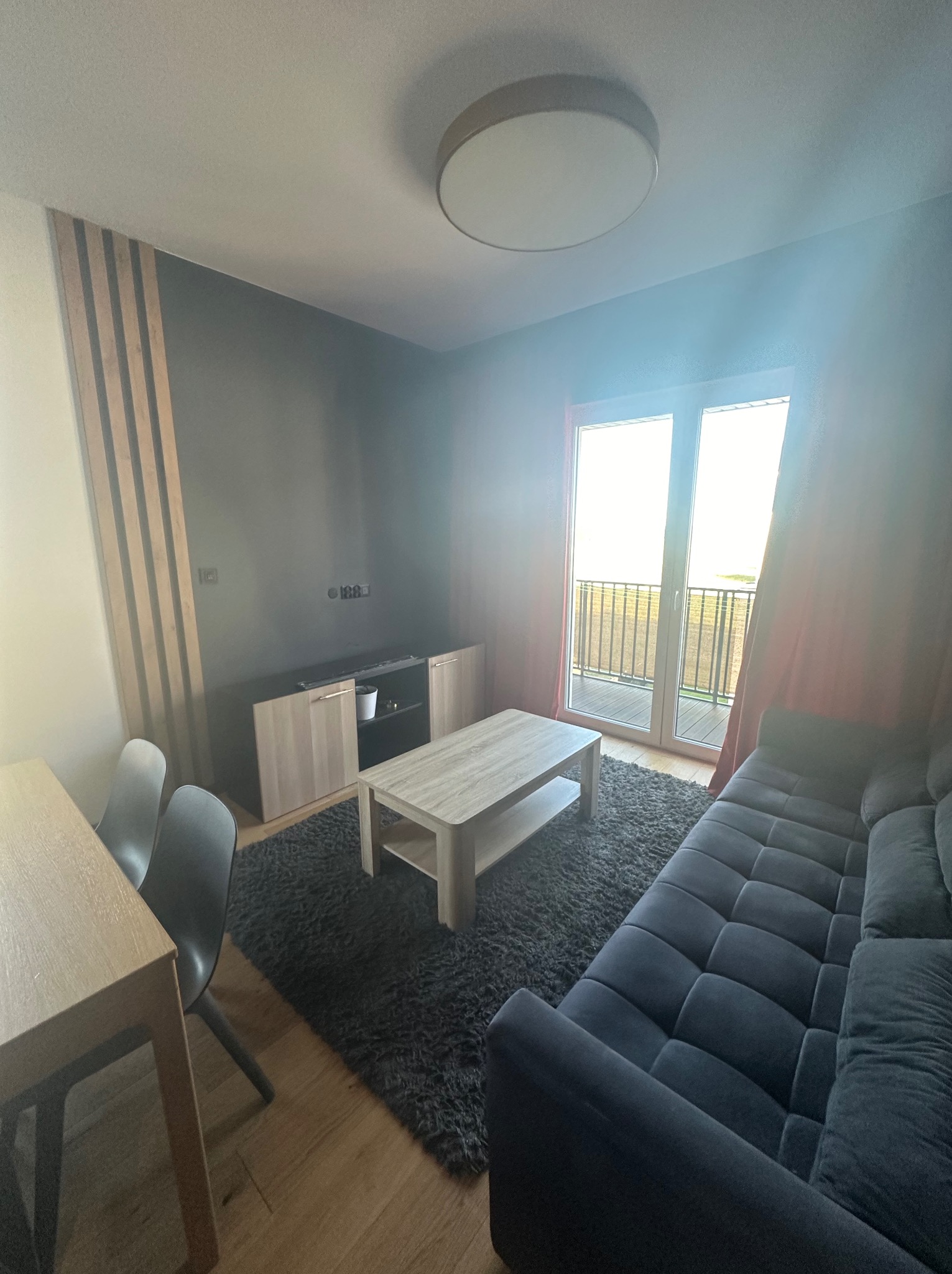 Nowoczesny salon z szarą ścianą akcentową, drewnianymi lamelami, ciemnoszarą sofą modułową, jasnym stolikiem kawowym na puszystym dywanie i wyjściem na balkon.