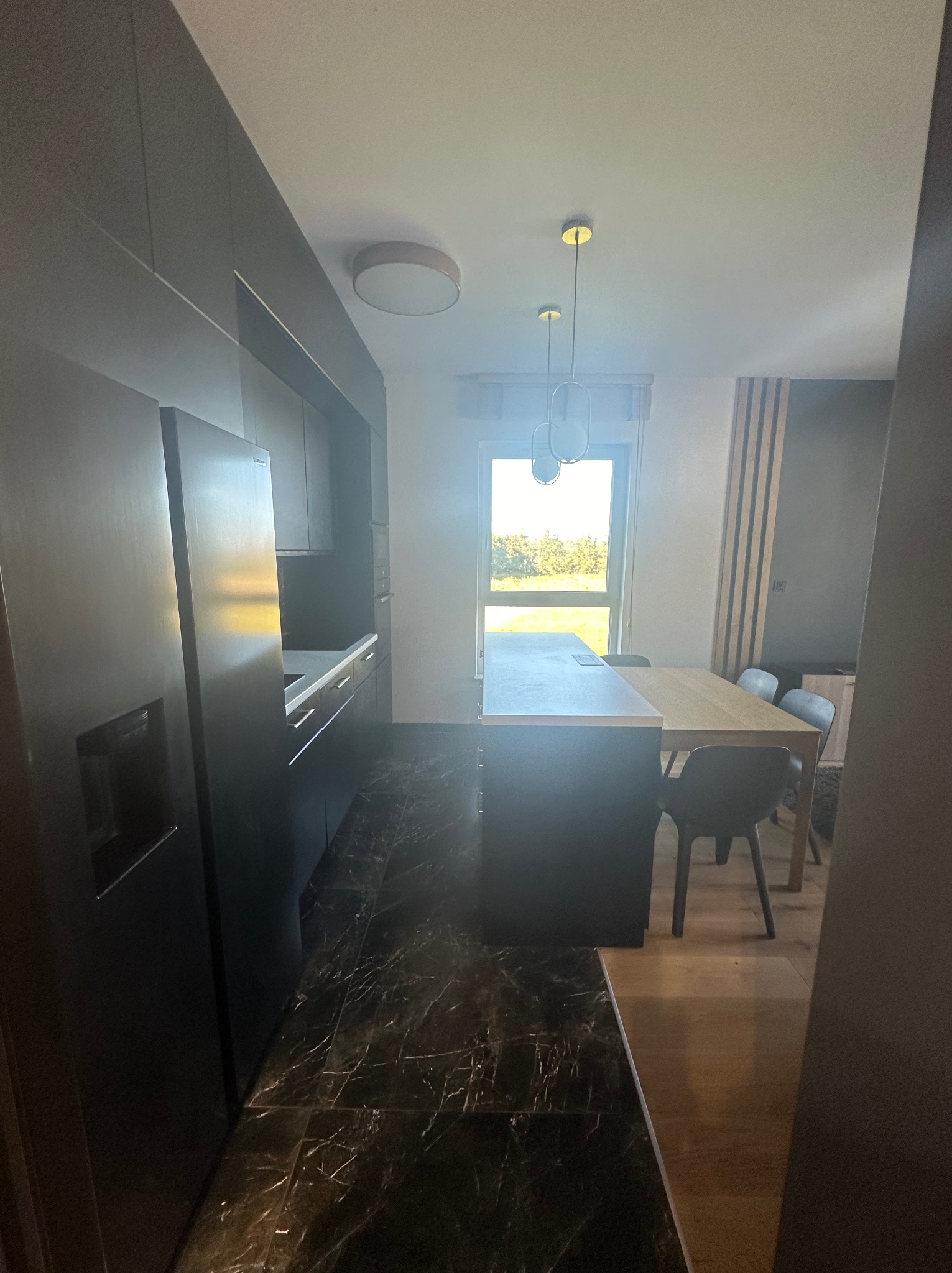 Nowoczesna, ciemna kuchnia z marmurową podłogą, oświetleniem punktowym i drewnianym stołem jadalnym przy oknie, widok z perspektywy wejścia.