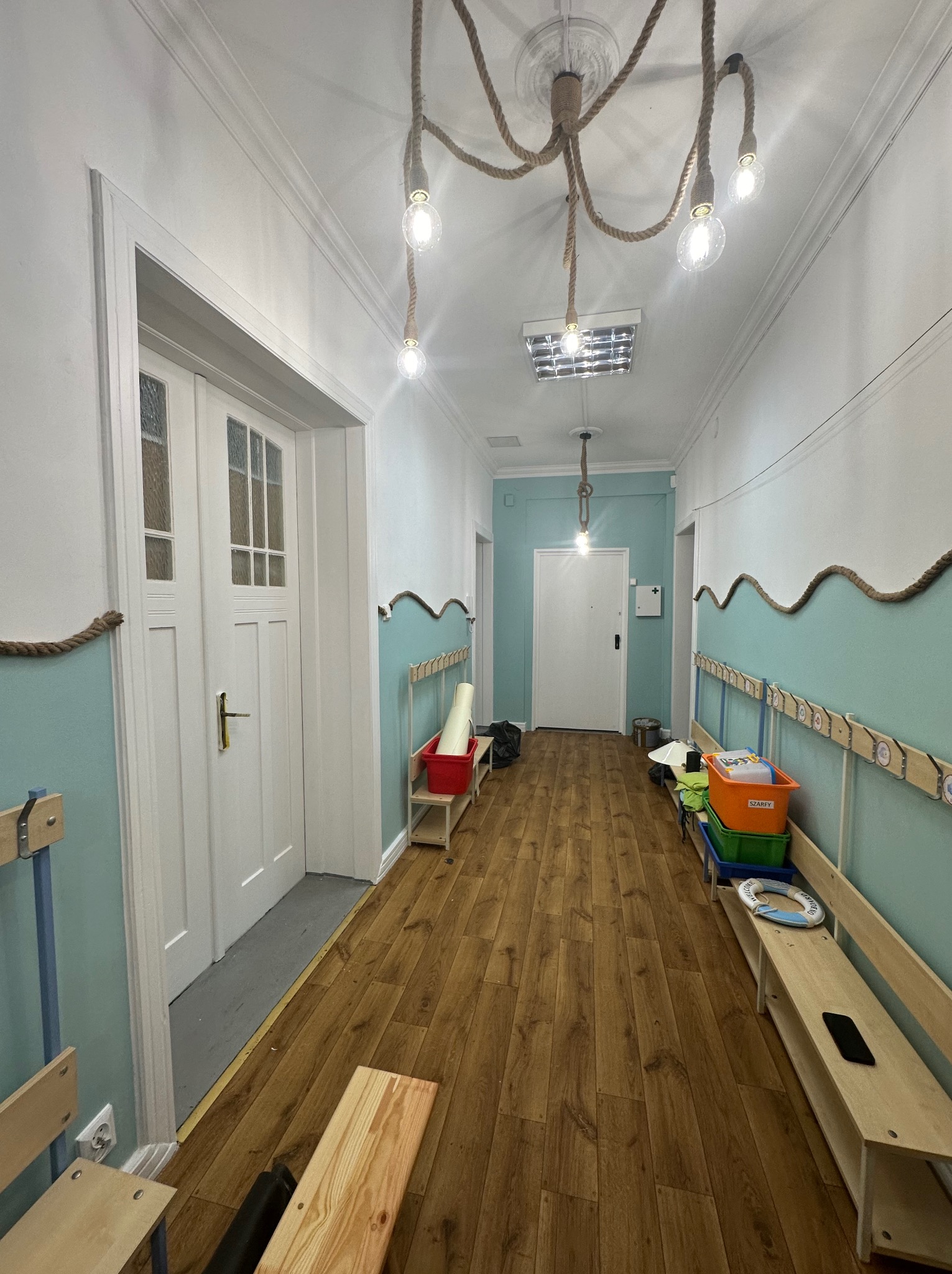 Korytarz w błękitno-białej kolorystyce z drewnianą podłogą i oryginalnym oświetleniem z liny jutowej, widoczne elementy dekoracyjne w stylu marynistycznym.