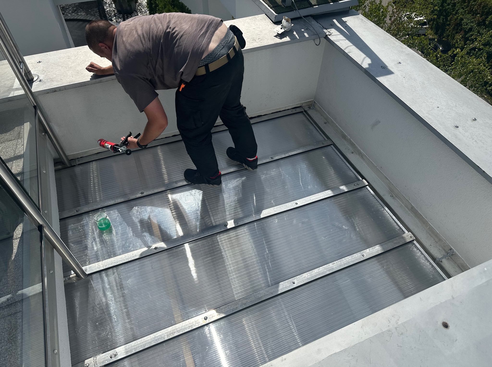 Pracownik uszczelnia silikonem połączenie paneli dachowych na tarasie, widoczny aplikator silikonu i pojemnik z płynem do czyszczenia.