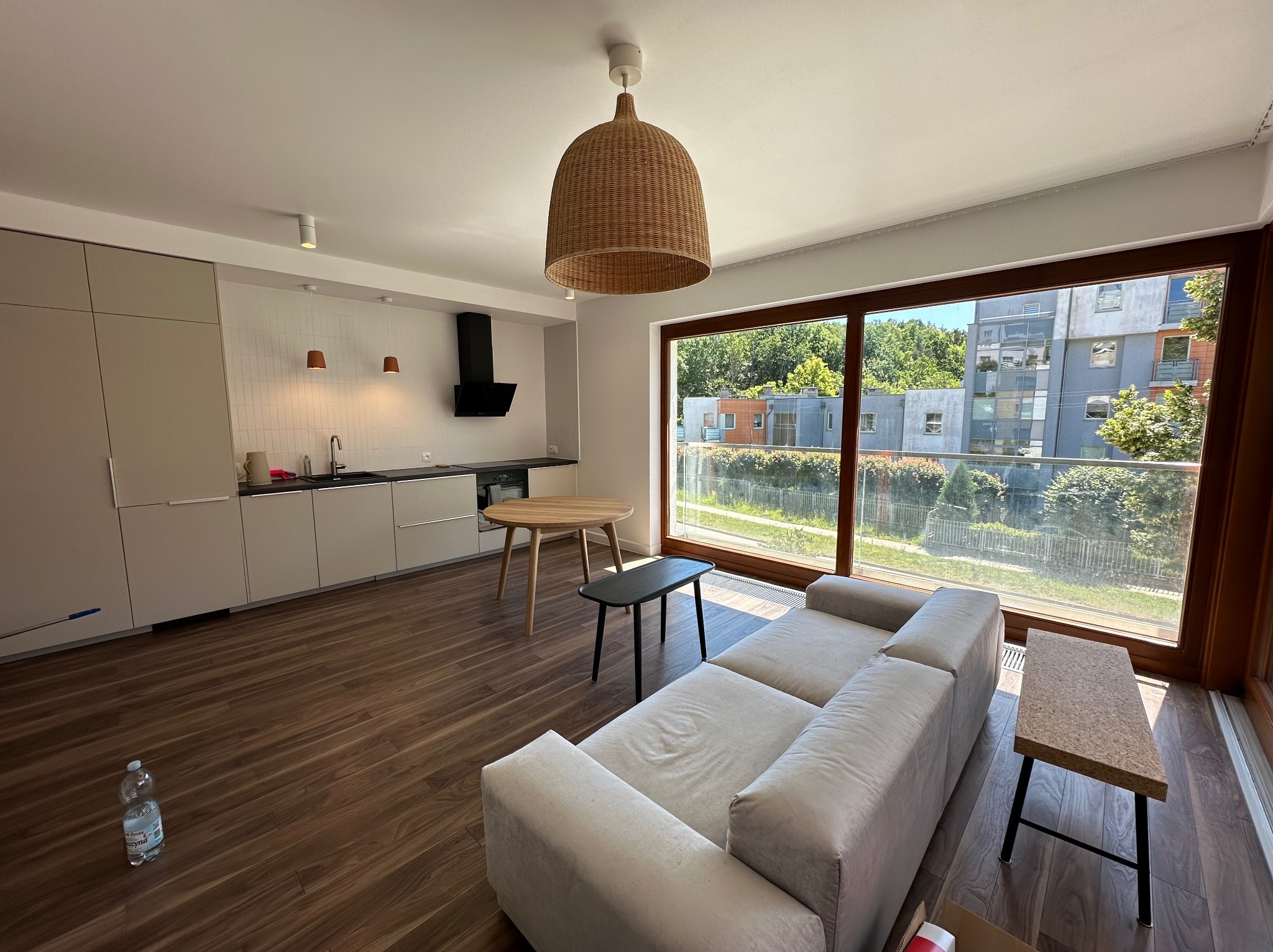 Jasne, przestronne wnętrze mieszkania po remoncie: nowoczesna kuchnia z białymi płytkami nad blatem, drewniana podłoga, duży narożnik i okno z widokiem na zieleń.