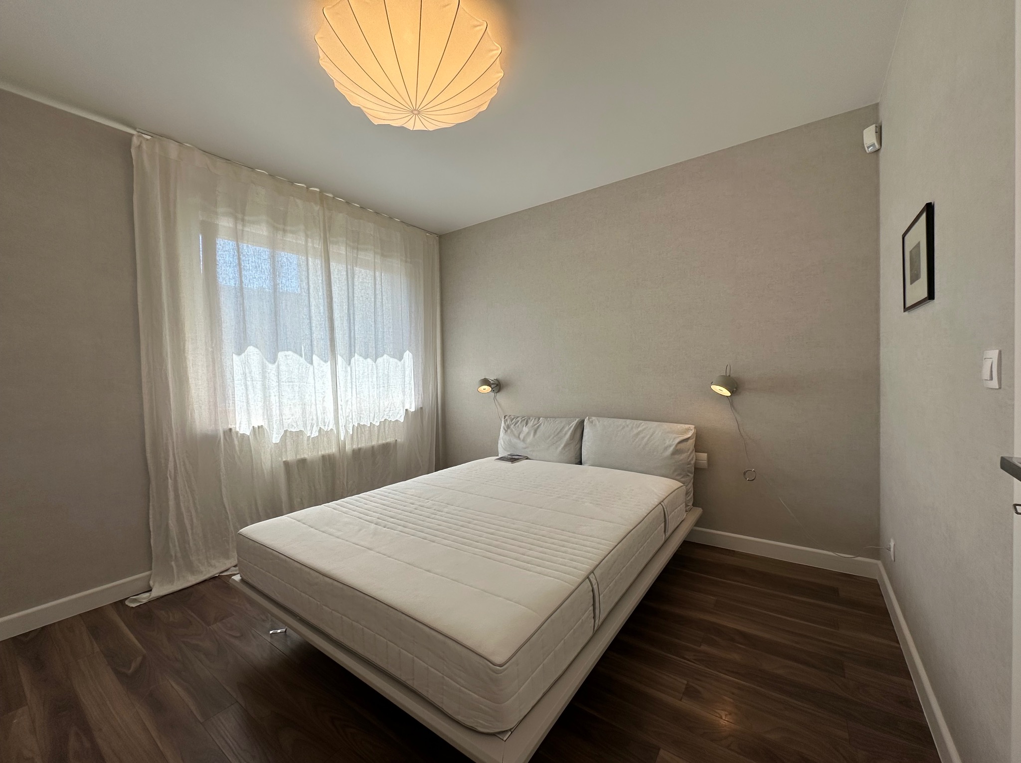 Sypialnia w Gdyni z drewnianą podłogą, beżowymi ścianami, białym łóżkiem z materacem i lampami ściennymi, oświetlona przez okno z delikatną, białą zasłoną.