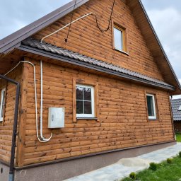 Zbigniew Drozdowski - Drewniany dom z brązowym dachem i widocznymi białymi rurami na elewacji, oświetlony naturalnym światłem, widziany z niskiej perspektywy.