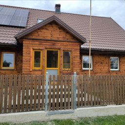 Zbigniew Drozdowski - Drewniany dom z brązowym dachem, panelami słonecznymi i żółtymi ramami okiennymi, widziany zza drewnianego płotu z metalowymi słupkami.