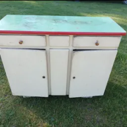 Stara, odrestaurowana szafka z zielonym blatem i czerwonym obramowaniem, stojąca na trawie, z otwartymi drzwiczkami ukazującymi wnętrze.