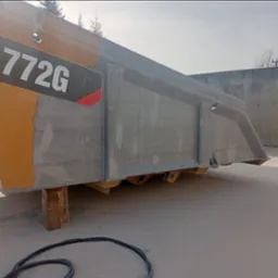 Fragment skrzyni ładunkowej ciężarówki 772G, w trakcie czyszczenia strumieniowo-ściernego, z widocznymi śladami piaskowania na żółtej powierzchni, na betonowym podłożu.