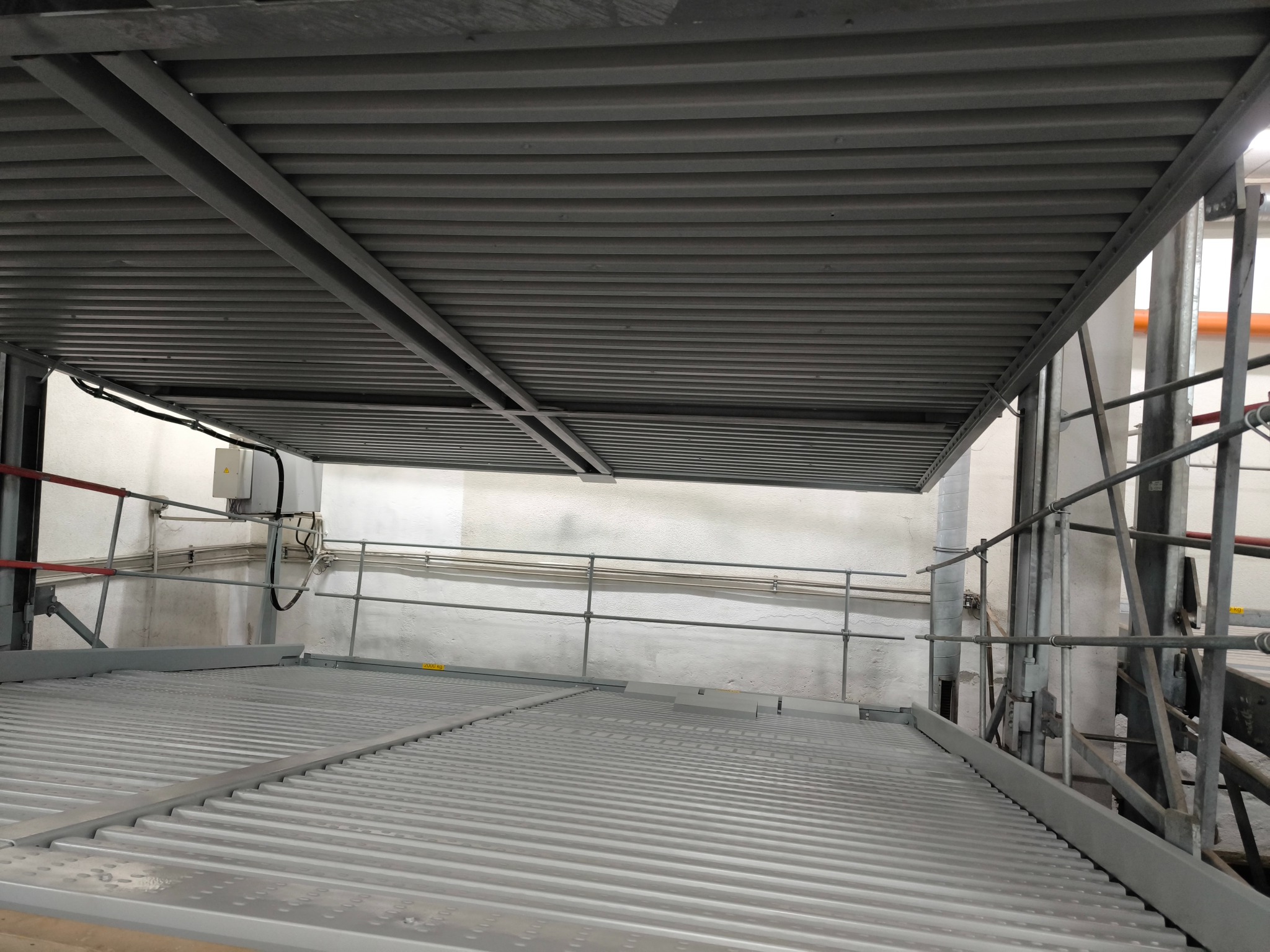 Metalowa konstrukcja sufitowa i podłogowa w hali przemysłowej z widocznymi rurami i kablami, pomalowana na szaro.