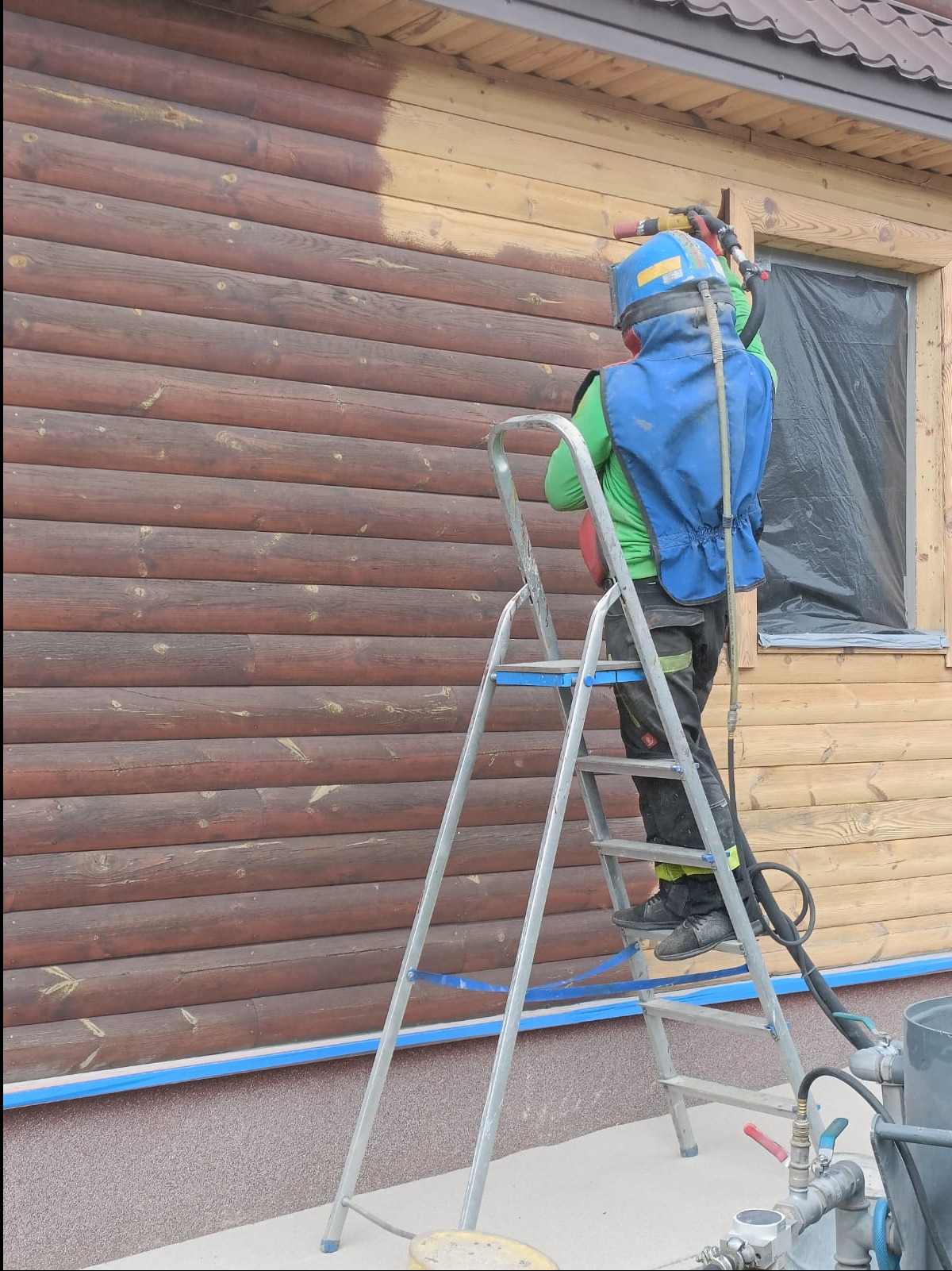 Pracownik w kombinezonie ochronnym stoi na drabinie i aplikuje preparat na drewnianą elewację domu, widoczny fragment elewacji po zabiegu i przed zabiegiem, okno zabezpieczone folią.
