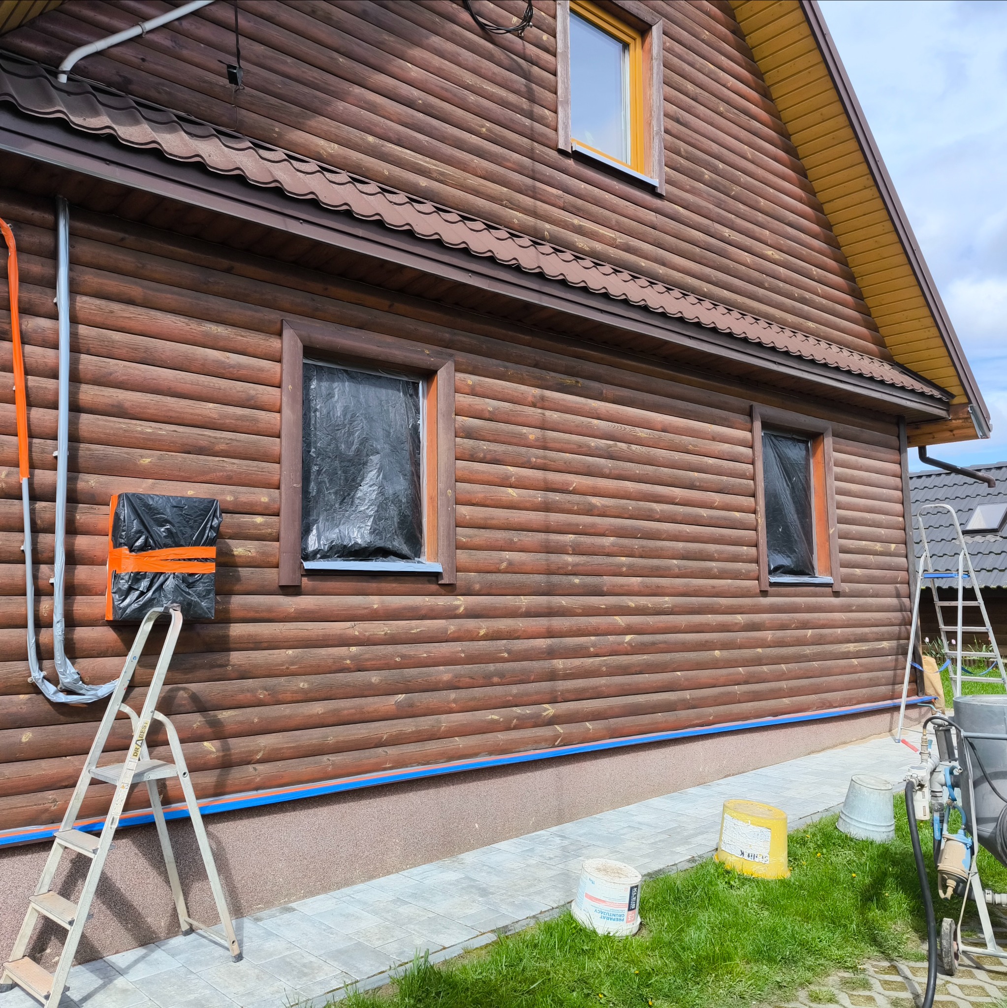 Przygotowanie drewnianej elewacji do malowania: okna zakryte folią, zabezpieczone elementy, drabina przystawiona do ściany, widoczne puszki z farbą i urządzenie do malowania natryskowego.