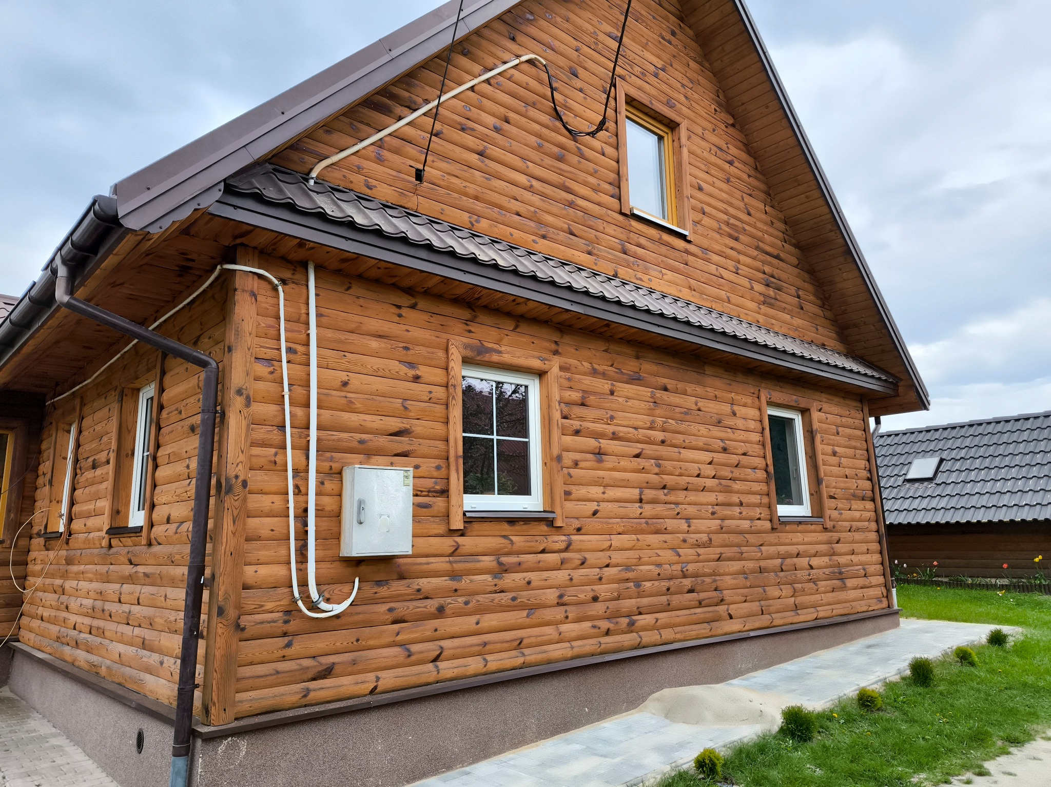 Drewniany dom z brązowym dachem i widocznymi białymi rurami na elewacji, oświetlony naturalnym światłem, widziany z niskiej perspektywy.