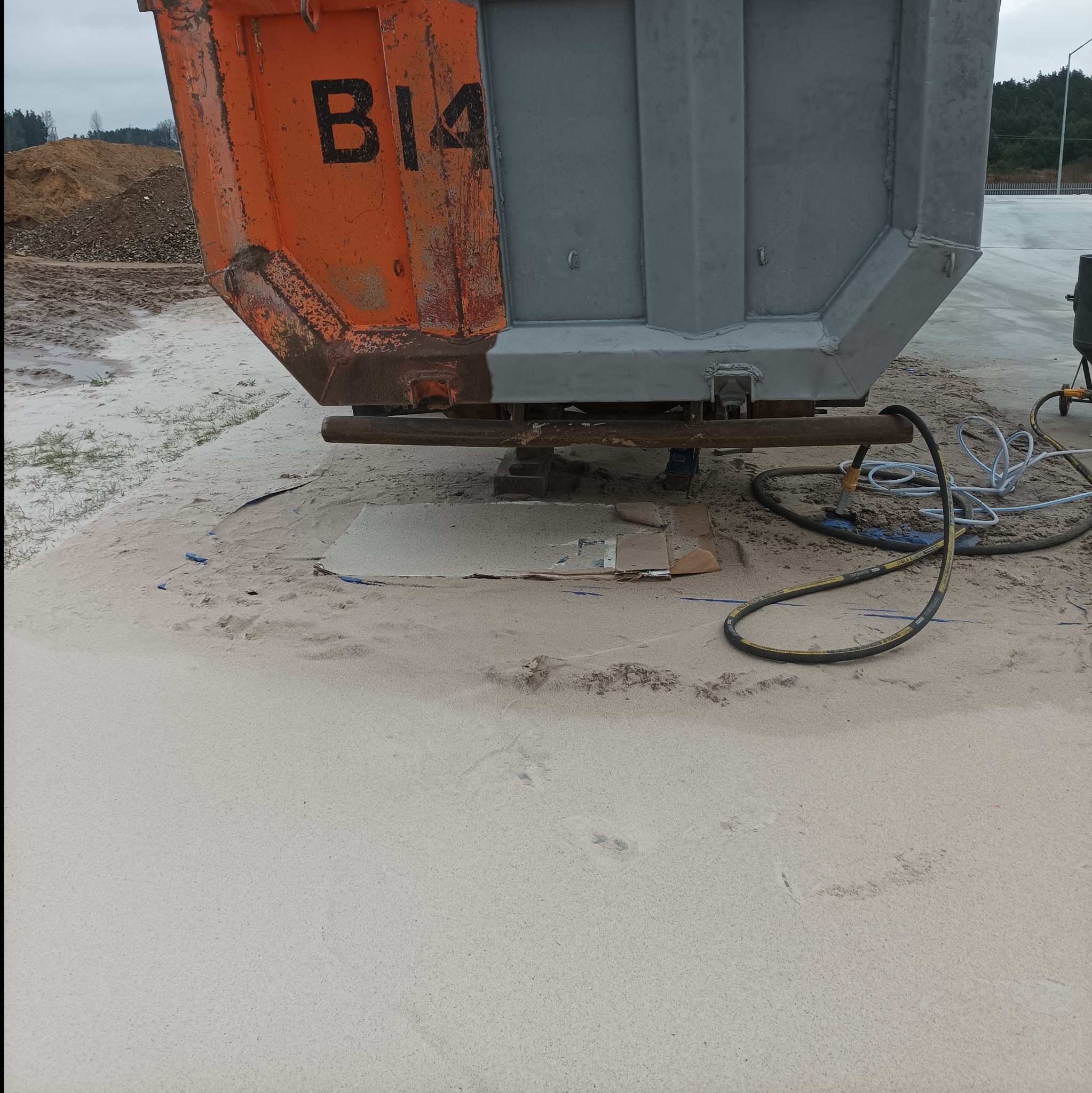 Kontener budowlany częściowo odnowiony, z widoczną różnicą między starą pomarańczową farbą z napisem B14 a nową szarą powłoką, stojący na piasku z widocznymi przewodami i elementami sprzętu...