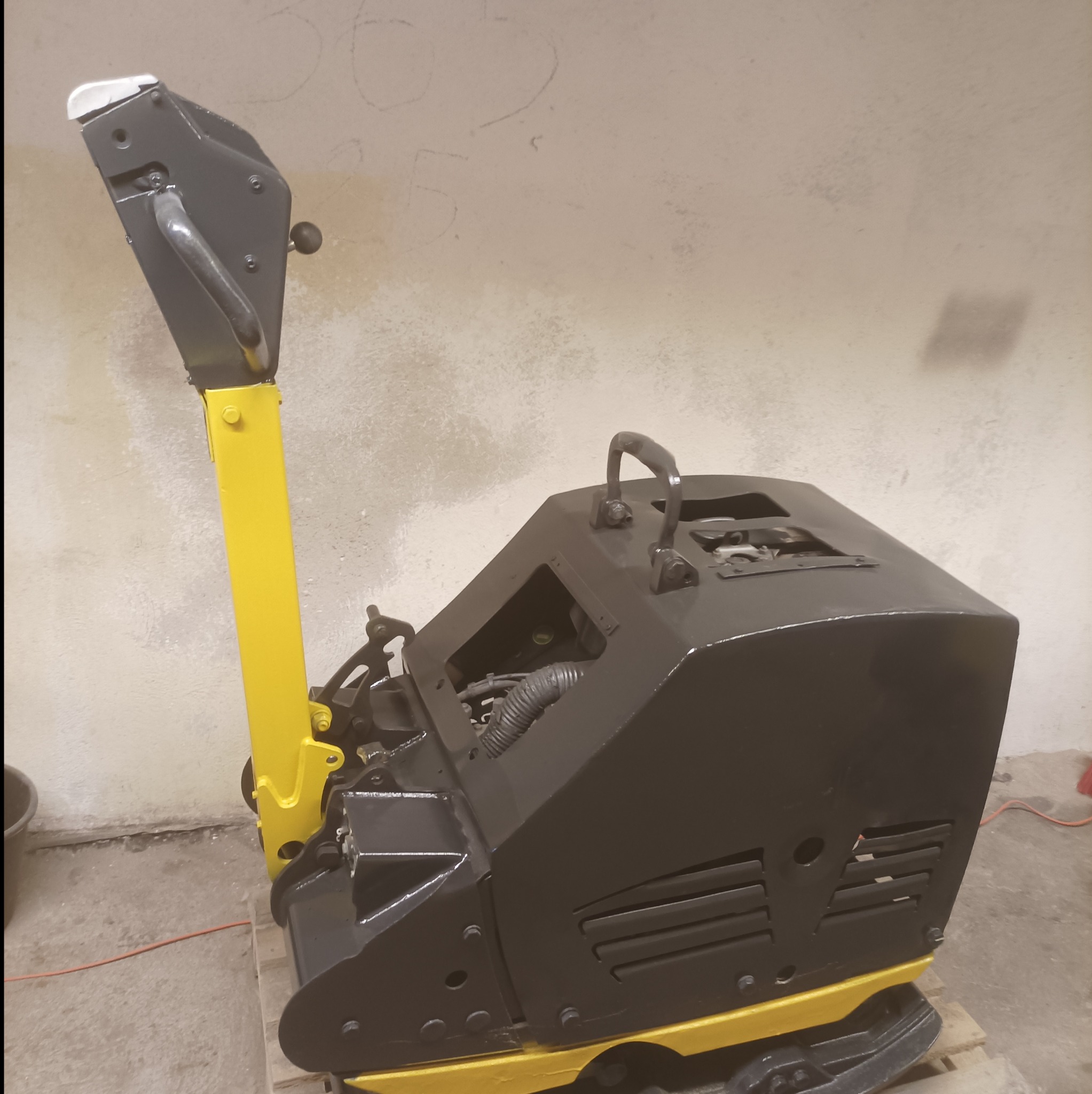 Zagęszczarka gruntu w kolorze czarnym z żółtymi elementami, stojąca na drewnianej palecie w pomieszczeniu z surową ścianą w tle.