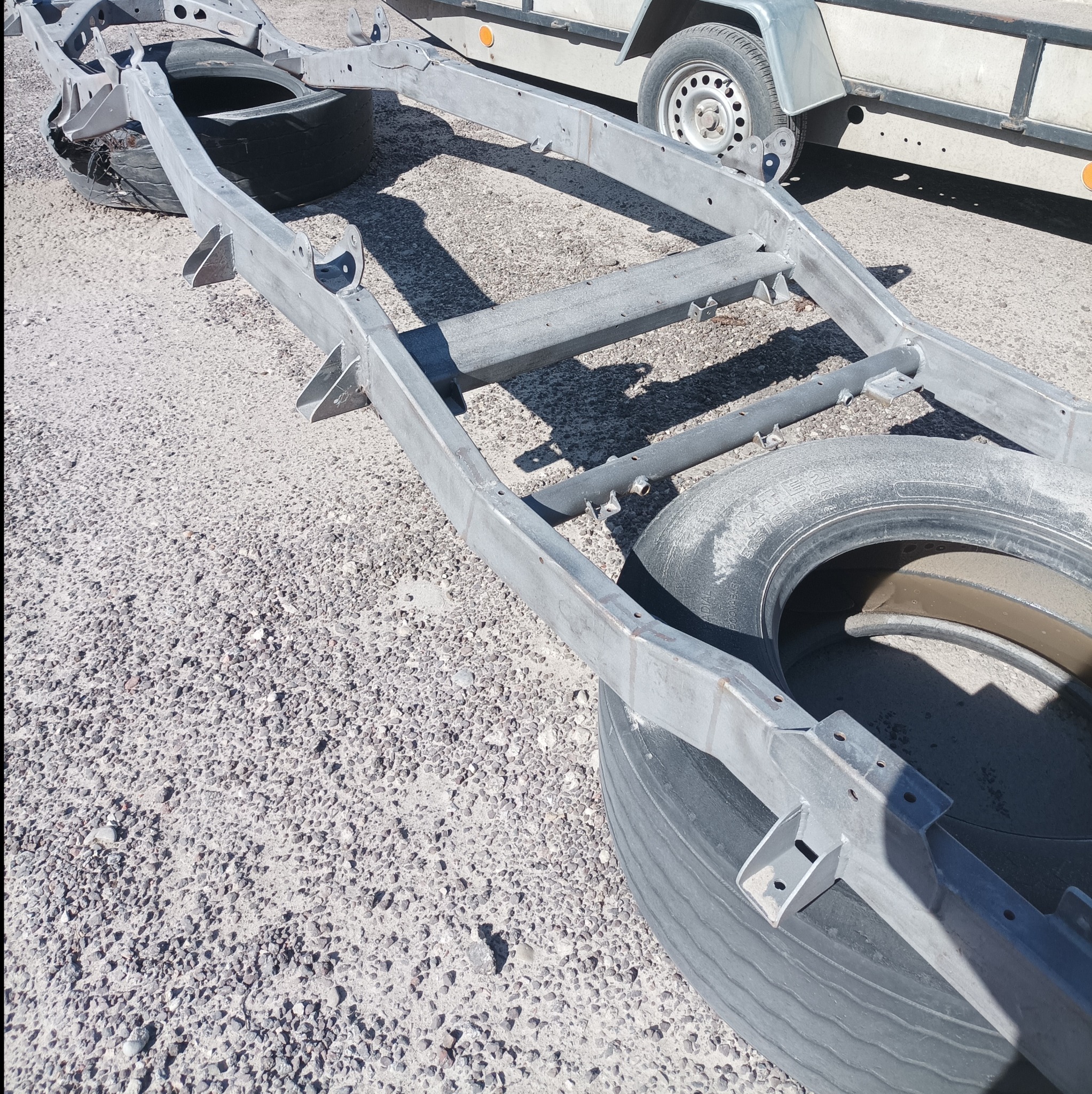 Oczyszczona rama pojazdu leżąca na oponach, widoczny fragment przyczepy w tle.